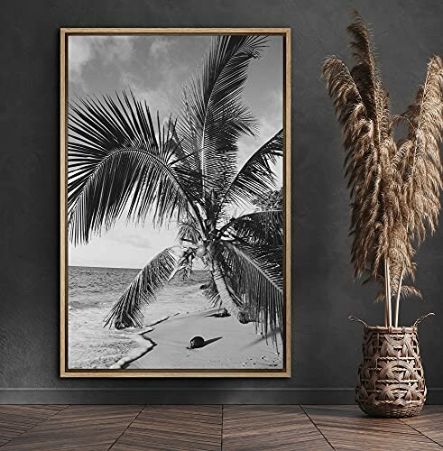 Black and white photo of a palm tree on a beach, framed and displayed on a wall next to a vase with decorative grasses.