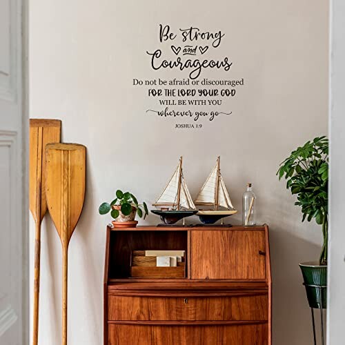 Wall decor with quote 'Be strong and courageous' above a wooden cabinet with boats and plants.